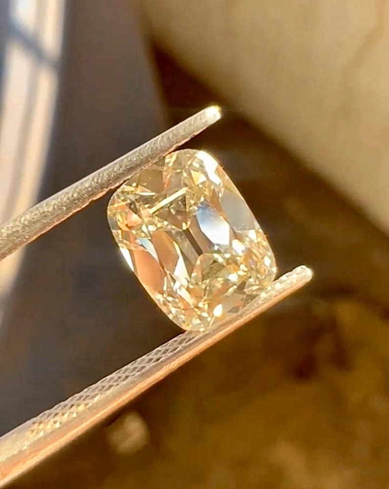K&ouml;nnte beinhalten: Ein kissenf&ouml;rmiger Diamant, gehalten von einer Pinzette. Der Diamant hat eine warme, gelbe Farbe und weist brillante Facetten auf. Die Pinzette ist silberfarben, der Hintergrund ist unscharf, um den Glanz des Edelsteins hervorzuheben.