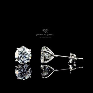 May include: A pair of round-cut stud earrings featuring clear, faceted stones set in silver-toned metal prongs. The text JEWELS BY JESSICA LAB GROWN DIAMONDS is centered above the earrings. The stones reflect light with a brilliant white sparkle.