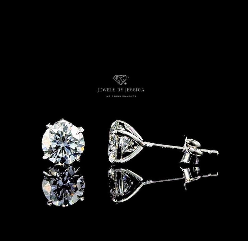 May include: A pair of round-cut stud earrings featuring clear, faceted stones set in silver-toned metal prongs. The text JEWELS BY JESSICA LAB GROWN DIAMONDS is centered above the earrings. The stones reflect light with a brilliant white sparkle.