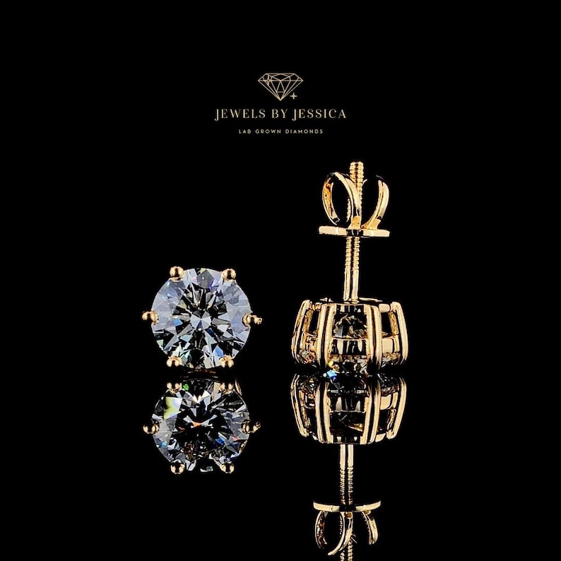 May include: Two gold stud earrings with round, clear lab-grown diamonds. The earrings have a simple design with a small, round diamond set in a gold setting. The earrings are displayed on a black background. JEWELS BY JESSICA LAB GROWN DIAMONDS is written in white text at the top of the image.