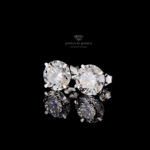 May include: A pair of round-cut stud earrings featuring clear, faceted stones set in silver-toned metal prongs. Above the earrings, the text JEWELS BY JESSICA LAB GROWN DIAMONDS is displayed with a small diamond icon on a black background.