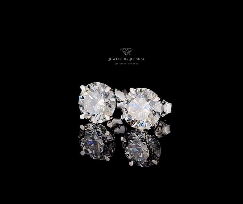 May include: A pair of round-cut stud earrings featuring clear, faceted stones set in silver-toned metal prongs. Above the earrings, the text JEWELS BY JESSICA LAB GROWN DIAMONDS is displayed with a small diamond icon on a black background.