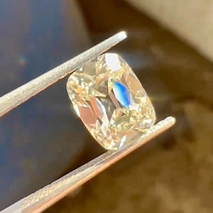 K&ouml;nnte beinhalten: Ein kissenf&ouml;rmiger Diamant, der von einer Pinzette gehalten wird. Der Diamant hat eine warme Champagnerfarbe mit Facetten, die das Licht reflektieren. Ein blauer Blitz ist im Stein sichtbar. Die Pinzette ist silberfarben.