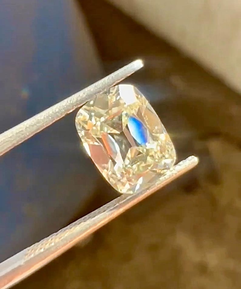 K&ouml;nnte beinhalten: Ein kissenf&ouml;rmiger Diamant, der von einer Pinzette gehalten wird. Der Diamant hat eine warme Champagnerfarbe mit Facetten, die das Licht reflektieren. Ein blauer Blitz ist im Stein sichtbar. Die Pinzette ist silberfarben.