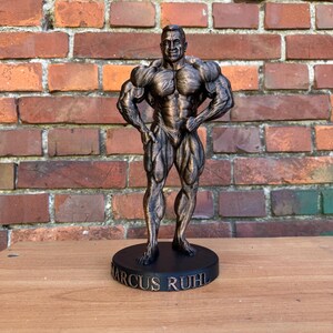 Può includere: Statua color bronzo di un bodybuilder in posa eretta, con le mani sui fianchi. La statua è su una base circolare nera con il nome "MARCUS RUHL" in lettere bianche. Sfondo: muro di mattoni.