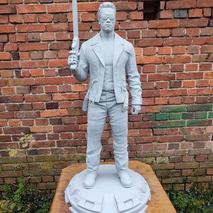 May include: A gray, unpainted, plastic figurine of a man wearing a leather jacket, jeans, and sunglasses. The figurine is holding a gun in his right hand and is standing on a round base.