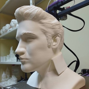 May include: A white 3D printed bust of a person with a pompadour hairstyle. The bust is on a black surface.