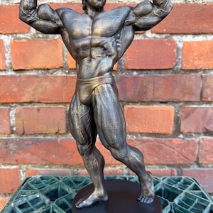 Könnte beinhalten: Eine bronzene 3D-gedruckte Statue von Arnold Schwarzenegger in einer Bodybuilding-Pose. Die Statue steht auf einem schwarzen Sockel mit der Aufschrift "ARNOLD".
