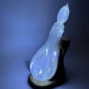 May include: A decorative, illuminated lamp shaped like a stylized gourd. The clear, textured body is lit with small white lights, casting a soft glow. The lamp sits on a dark brown base with a light brown trim. The overall design is organic and artistic.