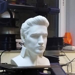 May include: A white 3D printed bust of a person with short hair. The bust is sitting on a black platform. The platform is on a 3D printer.