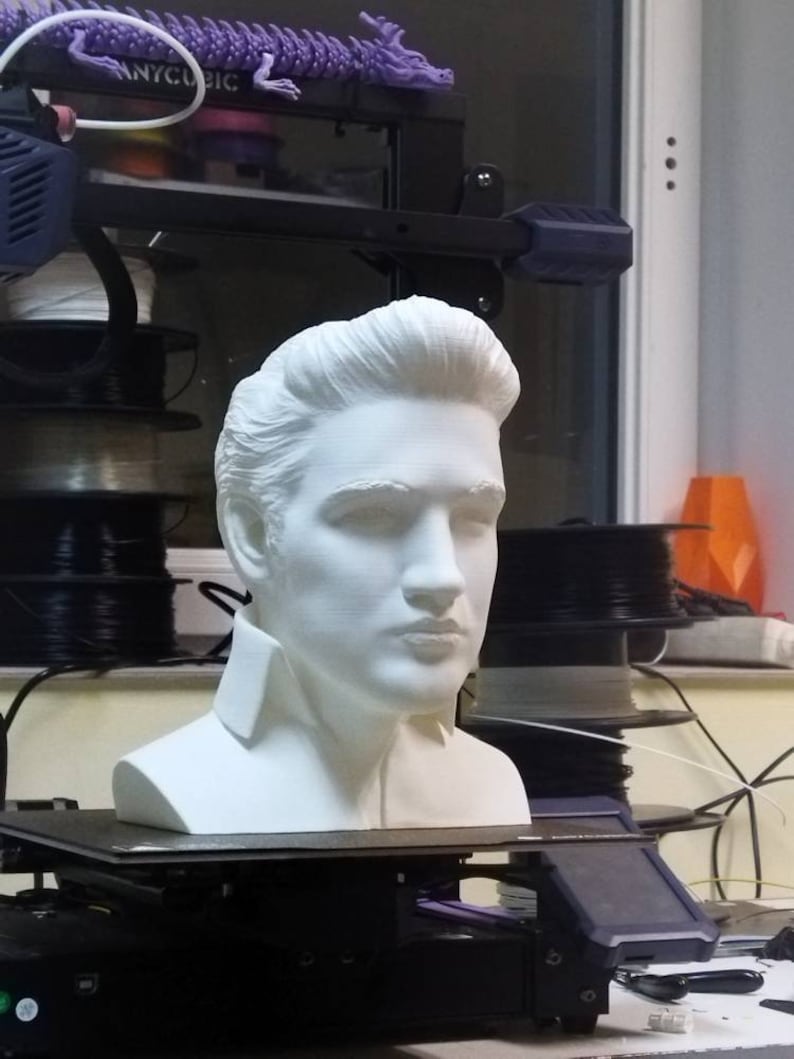 May include: A white 3D printed bust of a person with short hair. The bust is sitting on a black platform. The platform is on a 3D printer.