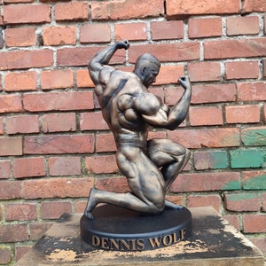 May include: Bronze-coloured statue of a muscular figure in a kneeling pose, with arms raised. The statue is on a black base with the name "DENNIS WOLF" inscribed in gold lettering.