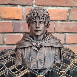Op de afbeelding: Een bronzen buste van een fictief personage met krullend haar, dat een bruin shirt en een mantel met een bladerenontwerp draagt. De buste staat op een zwart metalen rooster.