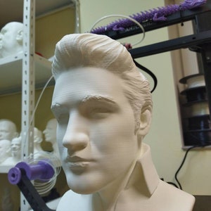 May include: A white 3D printed bust of a person with short, styled hair. The bust is on a black surface.