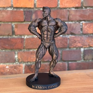 Könnte beinhalten: Eine bronzefarbene Statue eines Bodybuilders in klassischer Pose, stehend auf einem schwarzen, kreisförmigen Sockel. Die Statue ist sehr detailliert und zeigt die Muskeldefinition. Auf dem Sockel steht der Name "RAMON DINO".