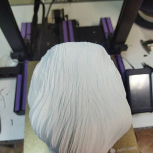 May include: A white 3D printed bust of a person with a textured surface. The bust is sitting on a gold surface with a black and white triangle logo.