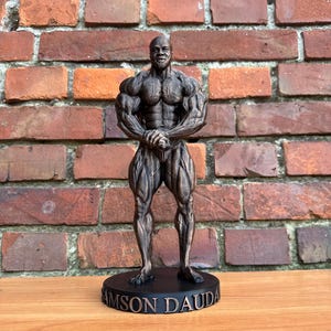 May include: A bronze-colored figurine of a muscular bodybuilder standing in a front double bicep pose. The figure is mounted on a circular black base with the text SAMSON DAUDA embossed in bronze lettering along the front edge of the base.