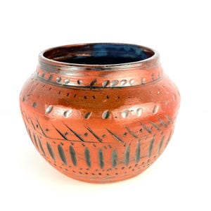 May include: A terracotta-coloured ceramic pot with a wide, rounded form. The pot has a dark blue interior and is decorated with etched patterns and dotted designs. The exterior has a glossy finish, highlighting the intricate details.