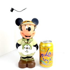 May include: A plastic figurine of Mickey Mouse dressed as a safari explorer. Mickey is wearing a khaki safari outfit, a brown hat, and a compass around her neck. She is holding a compass in her right hand. The figurine is about 12 inches tall and is standing next to a can of La Croix sparkling water.