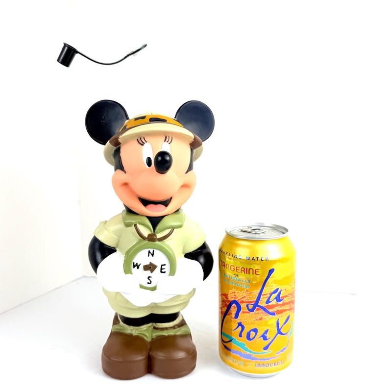 May include: A plastic figurine of Mickey Mouse dressed as a safari explorer. Mickey is wearing a khaki safari outfit, a brown hat, and a compass around her neck. She is holding a compass in her right hand. The figurine is about 12 inches tall and is standing next to a can of La Croix sparkling water.