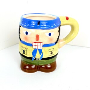 May include: A ceramic mug shaped like a boy scout with a blue bandana, yellow neckerchief, and a green uniform. The mug has a brown handle and the words "Boy Scouts of America" are printed on the side.