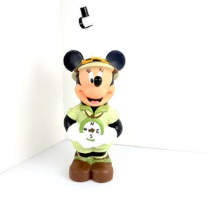 May include: A green and brown plastic figurine of Minnie Mouse dressed as an explorer. She is holding a compass with the letters N, W, E, and S.