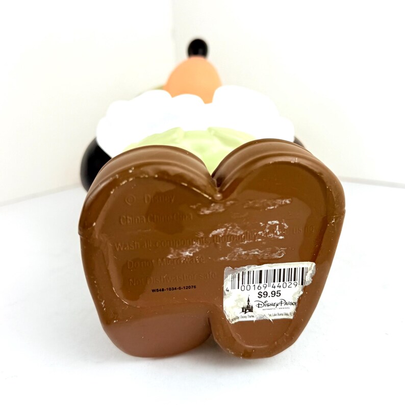 May include: A brown ceramic figurine of a cartoon character wearing a green shirt, white collar, and black pants. The figurine is standing on a brown base with a heart shape. The base has a label with the text "Disney", "Copyright Disney", "Wash with a sponge and mild detergent", "Do not microwave", "Made in China", "W948-1034-6-1307H", "0016944029", "$9.95", and "Disney Parks".