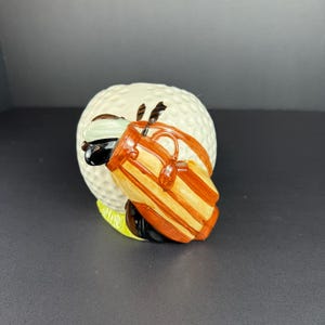 May include: A ceramic golf ball figurine with a golf bag. The golf ball is white with dimples, and the golf bag is brown with tan stripes. The figurine has a green base and black accents.