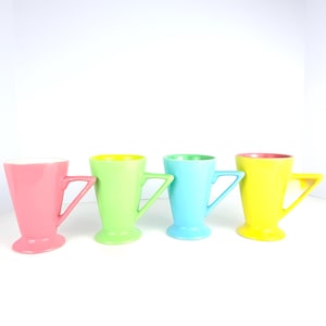 May include: Four colorful ceramic mugs in pink, green, blue, and yellow. Each mug has a unique handle design and a contrasting interior color. The mugs are arranged in a row against a white background.