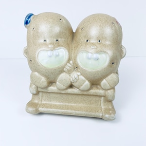 Puede incluir: Una figurita de cerámica de dos figuras sonrientes sentadas en un banco. Las figuras son beige con acentos azules y rojos.