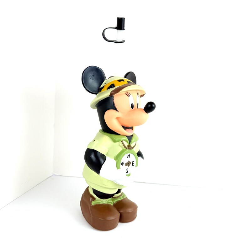 May include: A green and brown plastic figurine of Minnie Mouse dressed as an explorer. She is wearing a safari hat, a green shirt, and brown shorts. She is holding a compass with the letters N, W, E, and S.