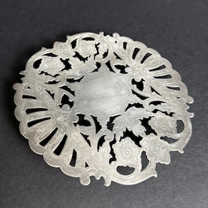 May include: A silver-colored, circular trivet with an intricate floral and leaf design. The trivet features a central, unadorned oval area surrounded by detailed cut-out patterns and floral motifs. The edges are scalloped, adding to its decorative appeal.