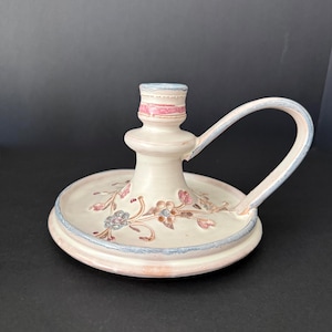 Puede incluir: Candelabro de cerámica color crema con asa. El candelabro tiene una base redonda y una columna central con un receptáculo para velas. Diseños florales y de hojas pintados en la base. El asa y los bordes están pintados con detalles azules y rosas.