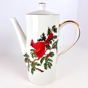 May include: White ceramic coffee pot with a red cardinal bird and holly berry design. The pot has a gold trim on the handle and lid. The spout is long and slender.