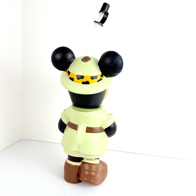 May include: A plastic figurine of Mickey Mouse dressed as an explorer. He is wearing a safari hat with a leopard print band, a khaki shirt and pants, and brown boots. He is standing on a white background.