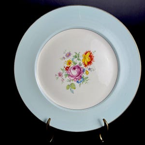 Puede incluir: Un plato redondo azul claro con borde dorado y diseño floral. El centro del plato es blanco con un ramo de flores rosas, amarillas y rojas. El plato se exhibe sobre un soporte dorado.