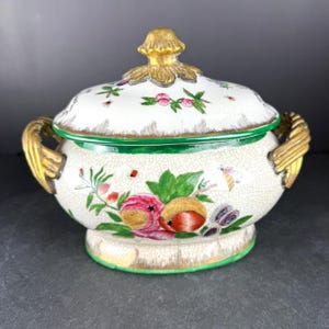 May include: A white ceramic lidded serving dish with a floral and fruit design. The dish has gold-colored handles and a gold finial on the lid. The rim is trimmed in green.