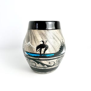 May include: A ceramic tumbler with a black rim and a wood-grain pattern. The tumbler features a black silhouette of a bird and a landscape design with a blue water line and black and grey decorative patterns.