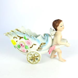 Könnte beinhalten: Vintage Keramik-Figur eines Cherubs, der einen dekorativen Wagen zieht. Der Wagen ist weiß mit blauen Akzenten, goldenen Verzierungen und floralen Details. Der Cherub hat braunes Haar, kleine Flügel und ein rosa Tuch.