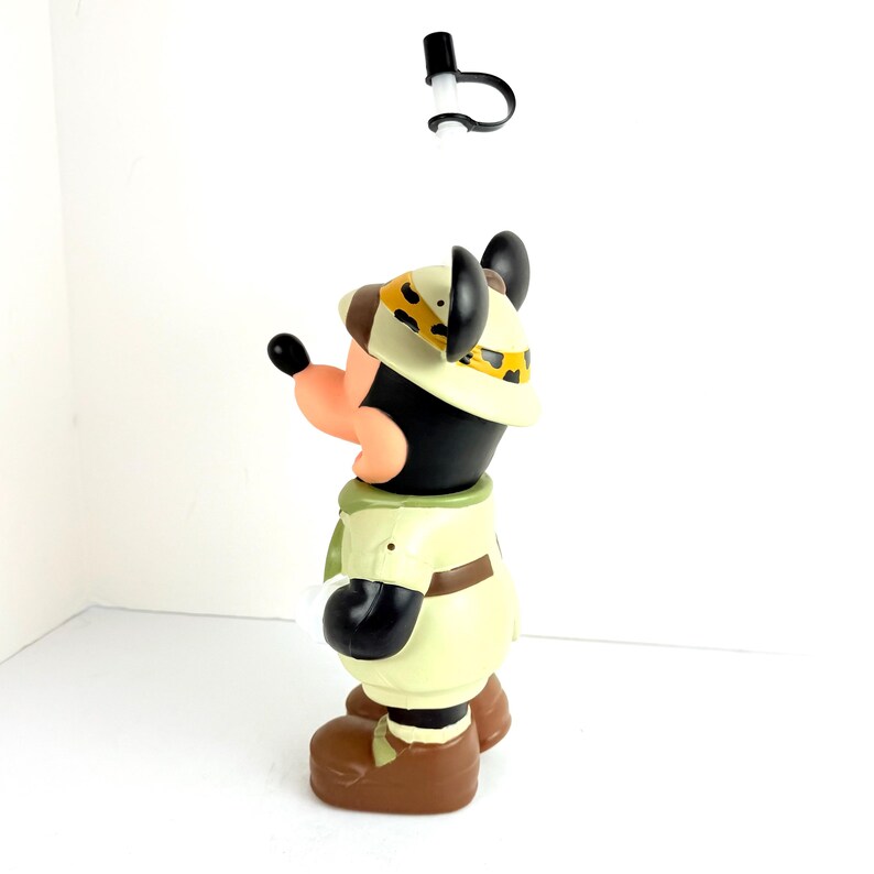 May include: A brown and black plastic figurine of Mickey Mouse dressed as an explorer. He is wearing a safari hat with a leopard print band, a khaki shirt, and brown pants. He is holding a straw in his mouth.