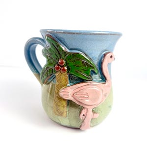 May include: A ceramic mug with a blue rim and handle, featuring a pink flamingo and a palm tree design. The mug has a green and tan base, with a blue sky background. The mug is approximately 4 inches tall.