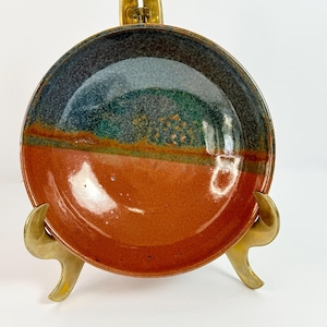 May include: A round ceramic bowl featuring a two-tone glaze design. The upper portion displays a mottled blend of dark blue, green, and grey tones, while the lower half is a solid, smooth terracotta orange. The bowl rests on a gold metal stand.