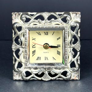 May include: A small, square quartz clock with a silver filigree frame. The clock face is cream-colored with black Roman numerals and hands. The word "QUARTZ" is printed below the center.