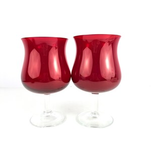 Peut inclure: Deux verres à vin rouge rubis, avec une coupe en forme de tulipe, une tige transparente et un pied transparent. Les verres sont côte à côte sur un fond blanc. Le matériau est brillant et réfléchissant.
