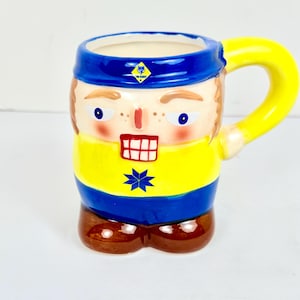 May include: A ceramic mug shaped like a person with a yellow, blue and brown design. The mug has a blue rim and a yellow handle. The person's face has a blue and white star on the chest. The mug has the text "The Scout" on the front.