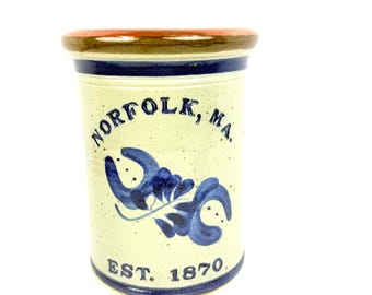 Puede incluir: Un recipiente cilíndrico de cerámica con un cuerpo color crema moteado. Presenta un diseño floral azul y el texto "NORFOLK, MA." y "EST. 1870." El borde es marrón y hay una banda azul en la parte superior e inferior.