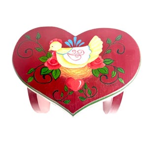 Puede incluir: Taburete de madera en forma de corazón, pintado de rojo con un diseño de gallina amarilla sobre un nido, rodeada de rosas rojas y hojas verdes. El taburete tiene tres patas rosas y un borde blanco.