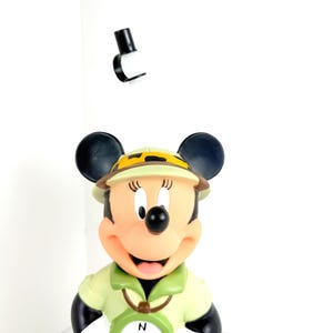 May include: A plastic figurine of Minnie Mouse dressed as an explorer. She is wearing a green safari shirt, a brown hat with a leopard print band, and a compass around her neck.