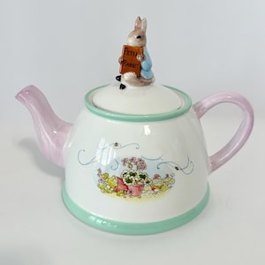 May include: A white ceramic teapot with a Peter Rabbit figurine on the lid. The teapot has a pink handle and spout, and a green trim. The body of the teapot features a floral design and the words "Peter Rabbit" on a sign.