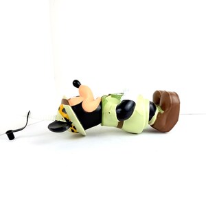 May include: A brown and black plastic figurine of Mickey Mouse dressed in a safari outfit. He is wearing a green and brown safari hat with a leopard print band, a green and brown safari shirt, and brown shorts. He is lying on his stomach with his head turned to the side. A black straw is attached to the figurine.
